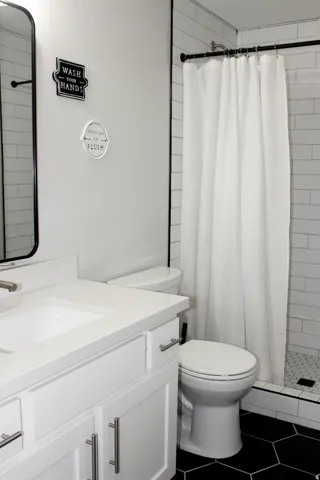 White vanity with a built-in sink and brushed nickel pulls, white subway tile shower surround, black hexagon floor tile, and a black-framed mirror