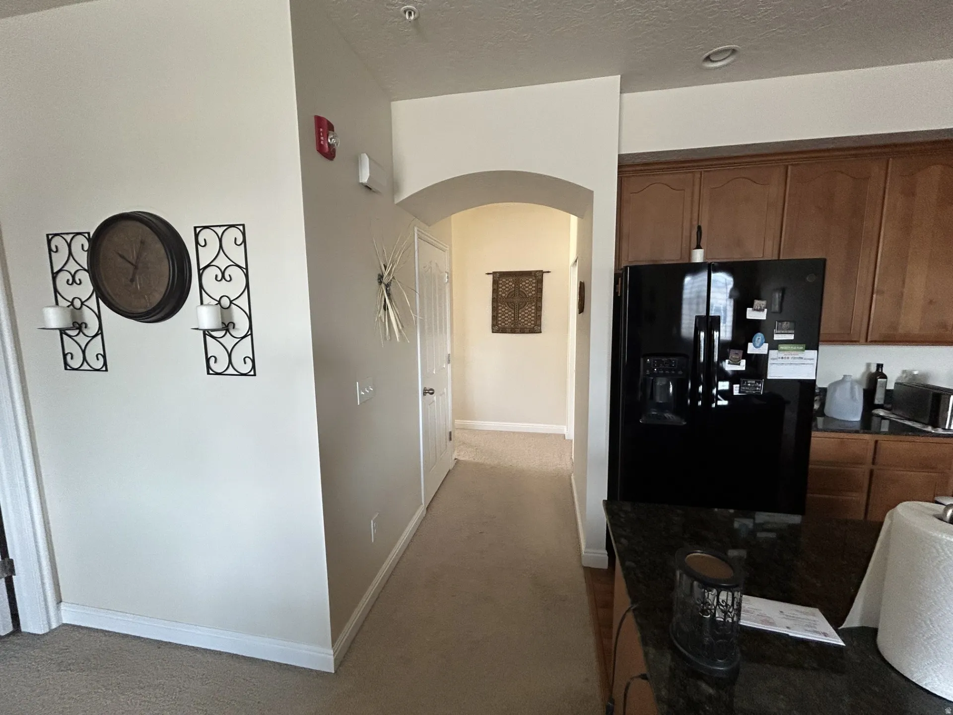Black refrigerator, granite-style countertops, wood-finish cabinetry, arched doorway & carpeted flooring.