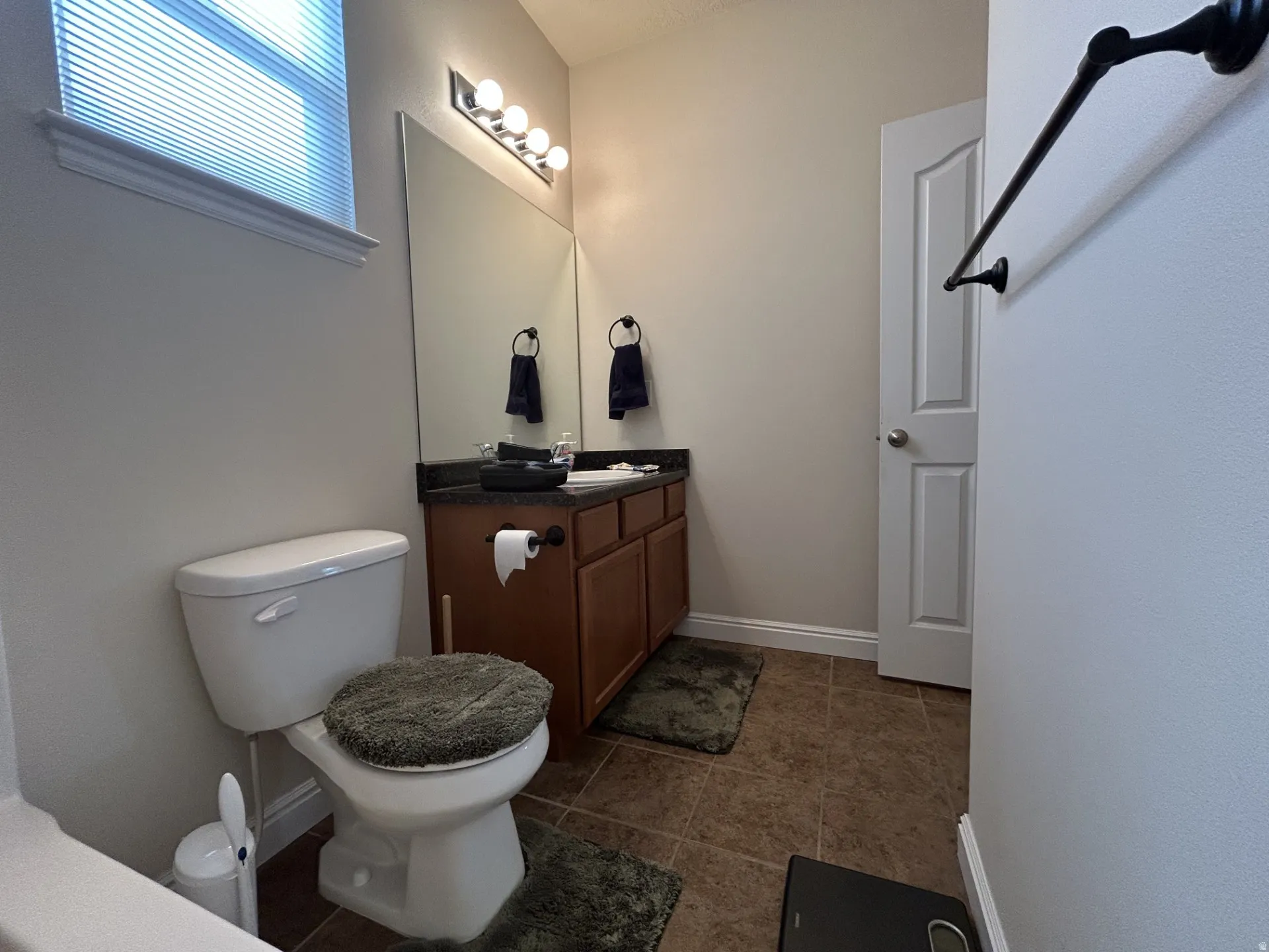 Bathroom featuring a single vanity with a dark countertop, white toilet, tile flooring & a window with blinds.