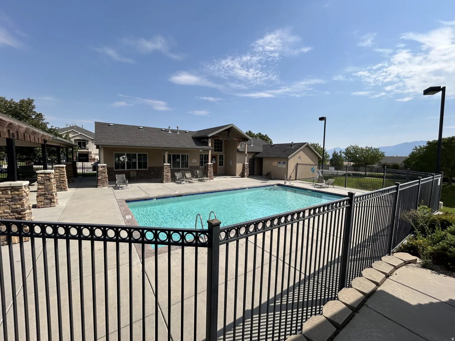 Outdoor swimming pool with surrounding concrete patio, black metal fencing, and an adjacent clubhouse/exercise building featuring stone accents