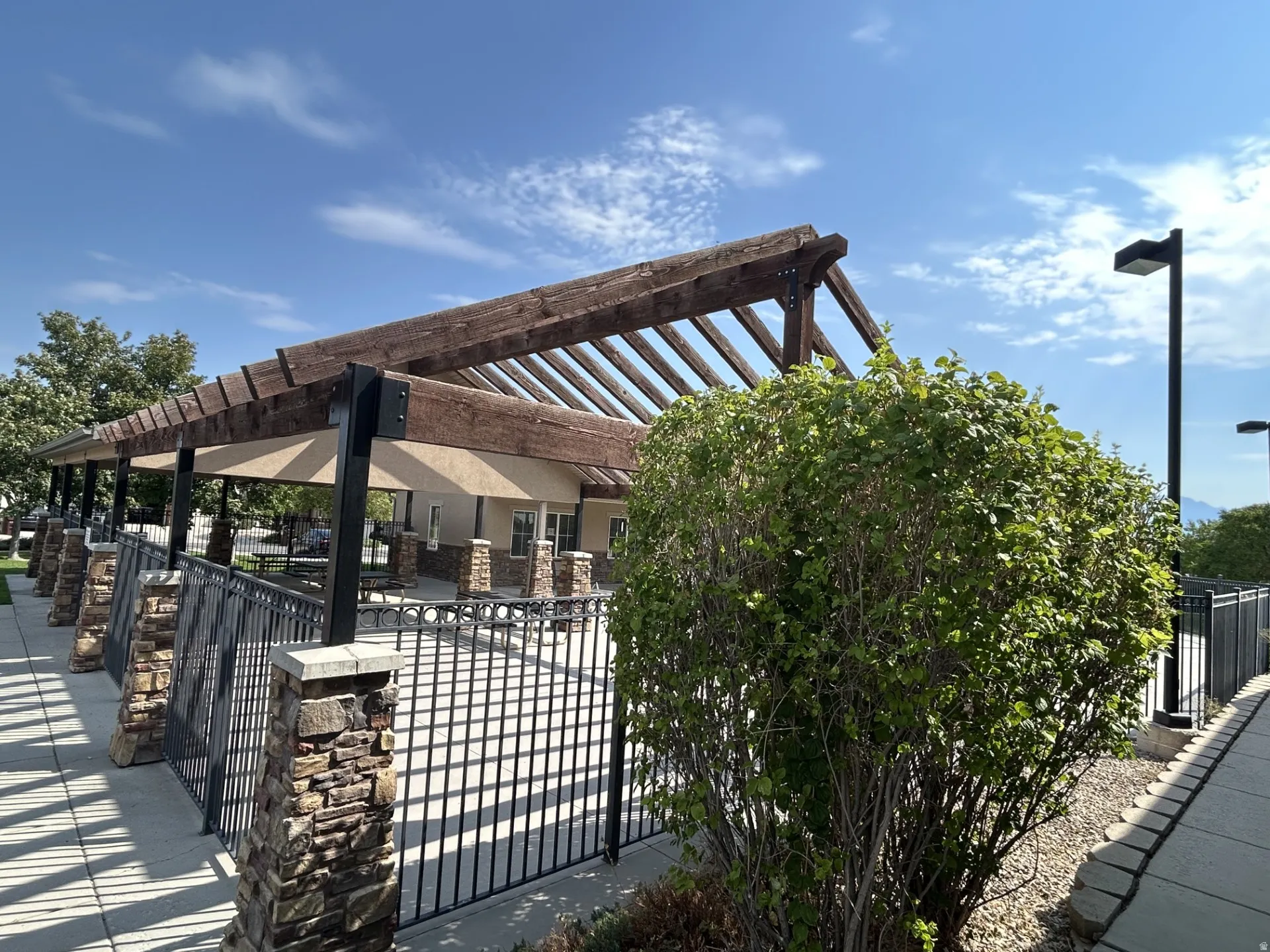 Wood pergola structure over patio, featuring stone-clad pillars & black metal fencing.