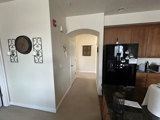 Black refrigerator, granite-style countertops, wood-finish cabinetry, arched doorway & carpeted flooring.