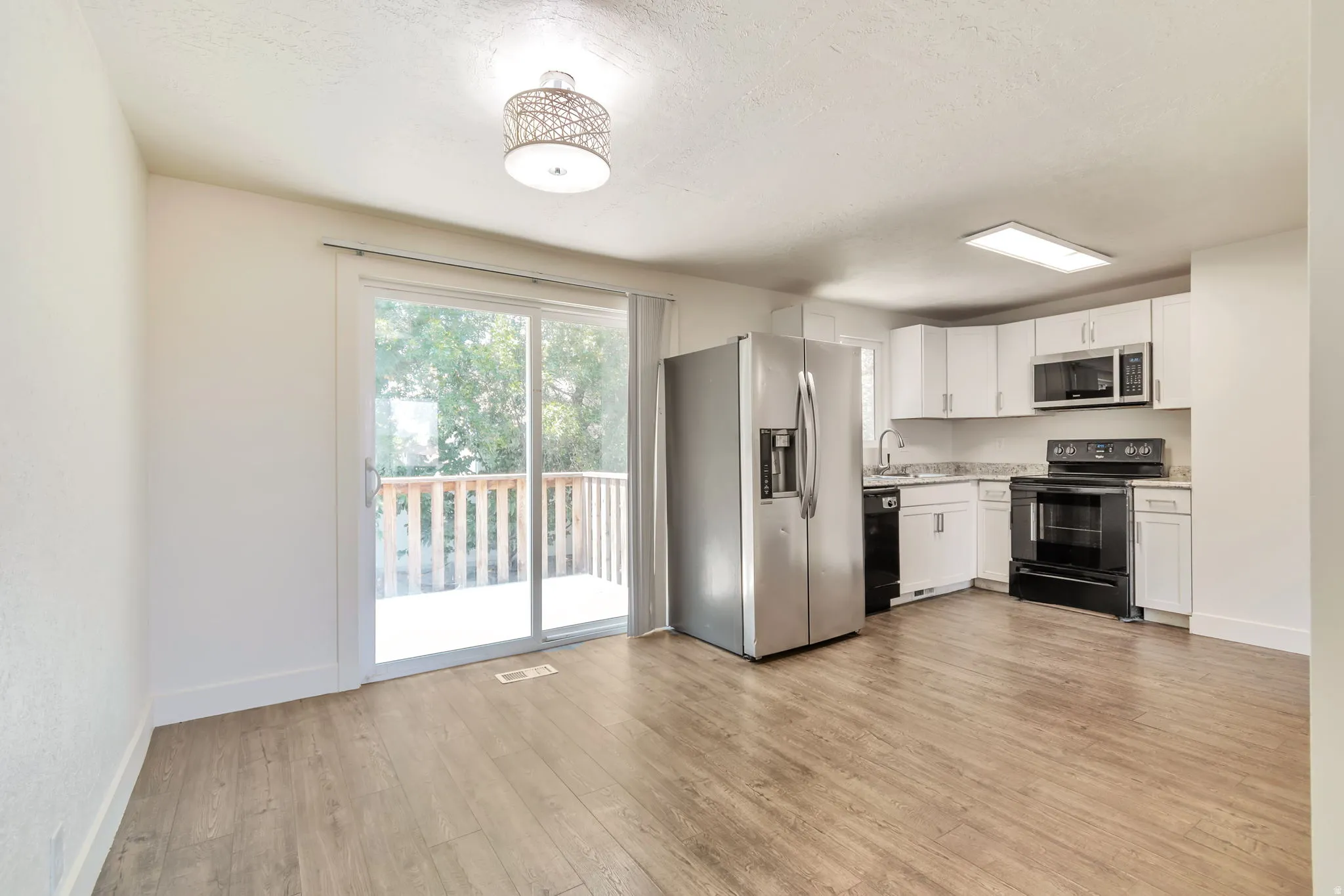 Open-concept living space with wood-finish flooring, white cabinetry, stainless steel appliances, and a sliding glass door leading to an outdoor deck