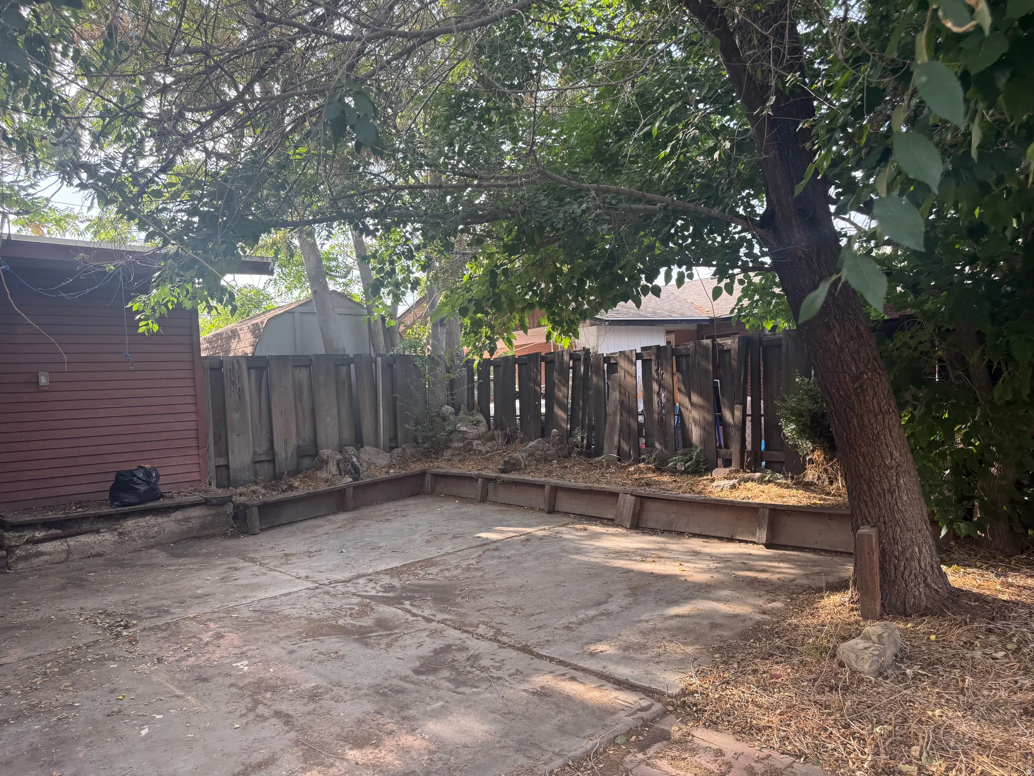 Concrete patio area with wood-framed raised beds, mature trees, and a wood fence enclosure