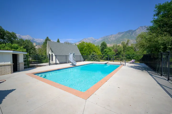 Outdoor swimming pool with concrete decking, surrounding black metal fencing, and a pool house featuring brick and horizontal siding