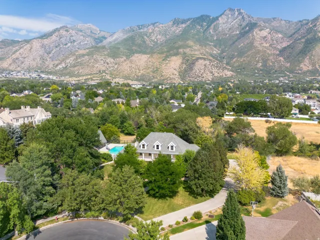 Traditional-style residence with a fenced swimming pool, surrounded by mature trees and a paved driveway, set against a mountainous backdrop