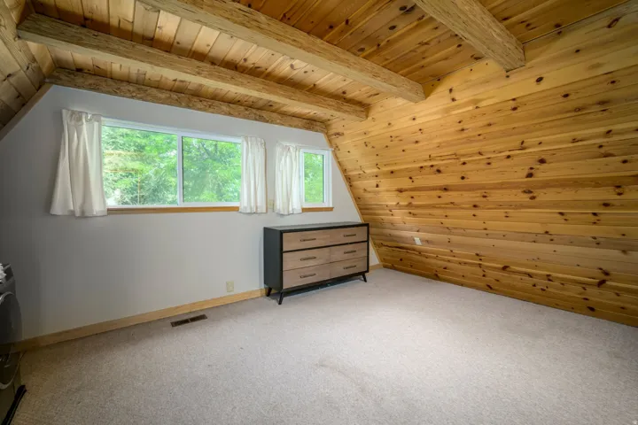 Exposed wood beams, sloped wood-paneled ceiling, light-colored carpeting, white window trim, and baseboards