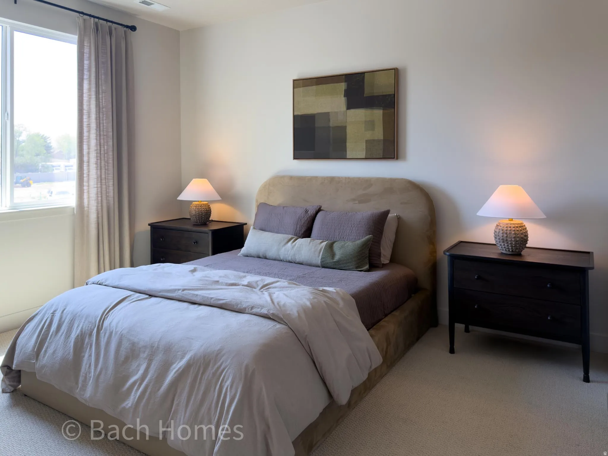 Master bedroom featuring an upholstered headboard bed with coordinating bedding, dual nightstands with table lamps, abstract wall art, and abundant natural light from a large window with thermal-lined curtains