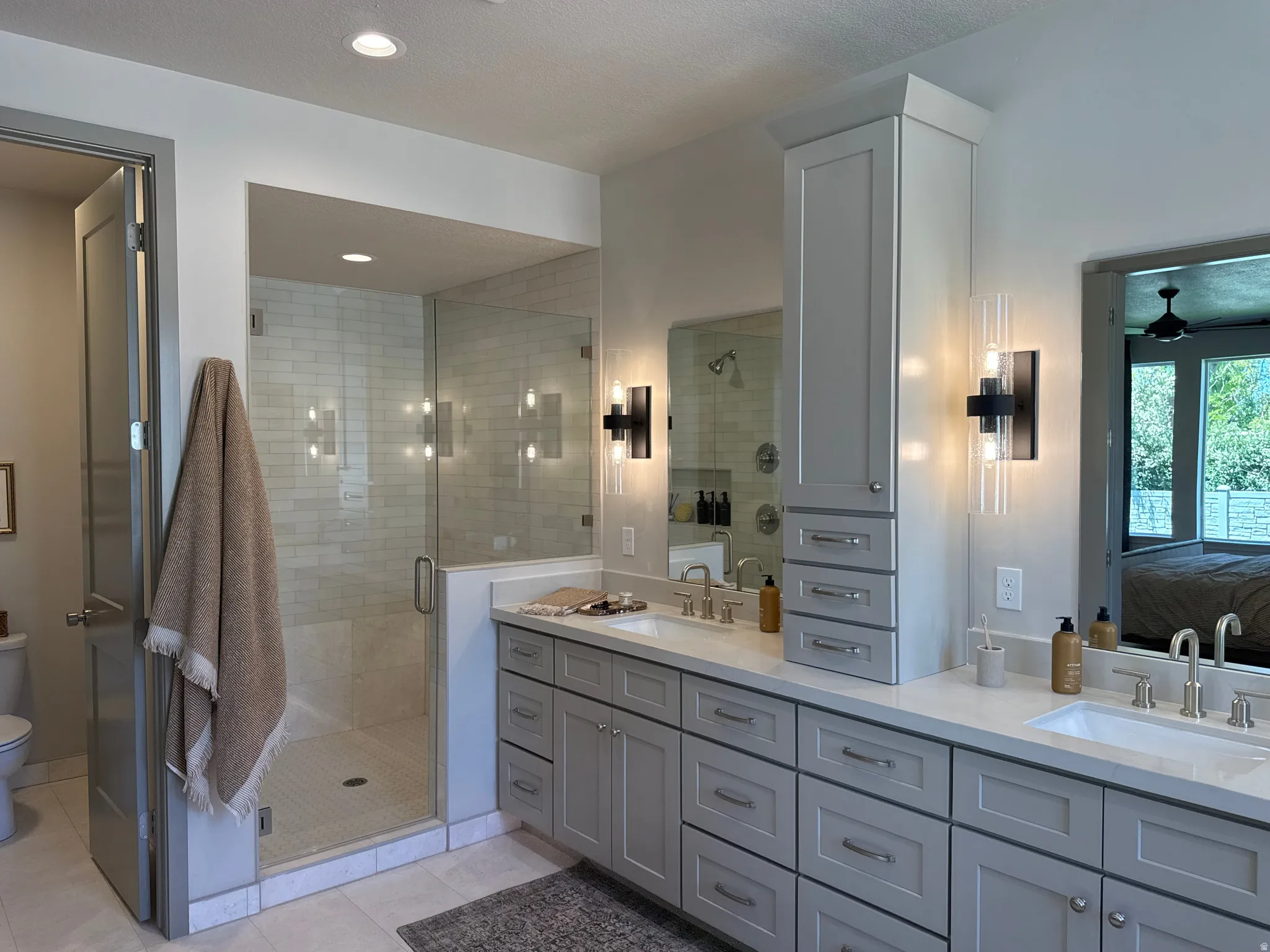Spacious bathroom featuring a frameless glass shower enclosure, double vanity with light gray cabinetry, built-in storage tower, light-toned countertops, and recessed lighting