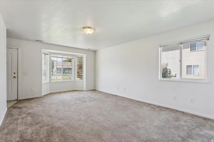 Bright, open living space featuring neutral carpet flooring, abundant natural light from multiple windows, recessed ceiling lighting, and clean white walls throughout