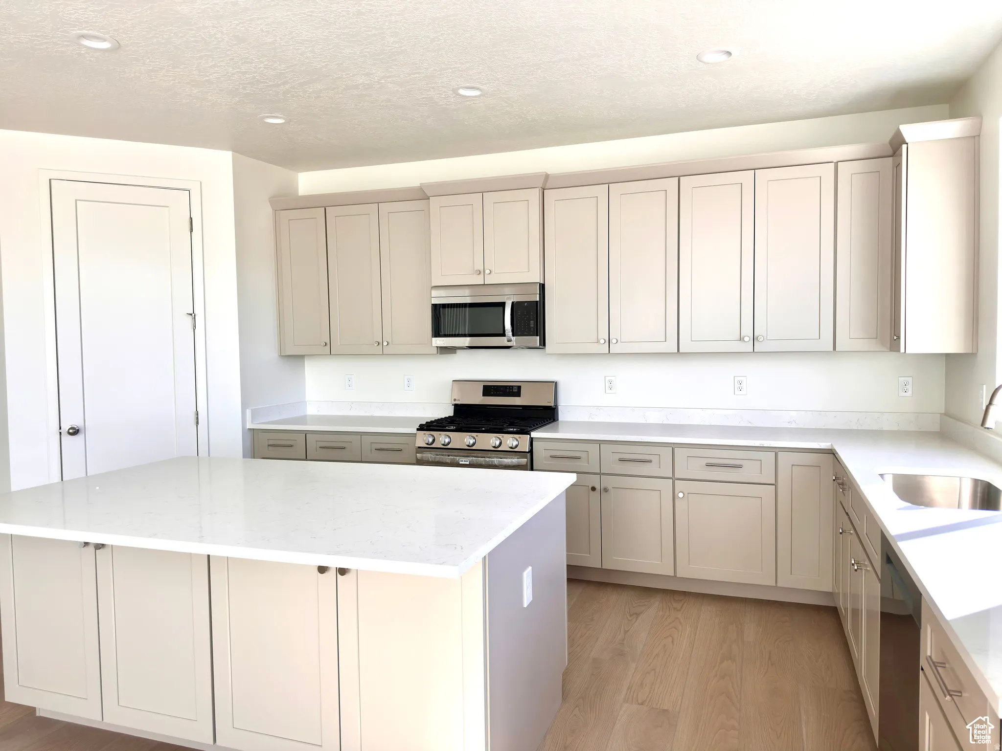 Bright kitchen featuring white shaker-style cabinetry, white quartz countertops, stainless steel appliances including a gas cooktop and built-in microwave, a large center island with seating, and wood-finish flooring throughout