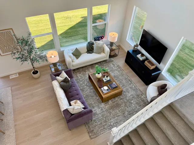 Spacious living area featuring light wood-finish flooring, multiple large windows, and a carpeted staircase with white railings