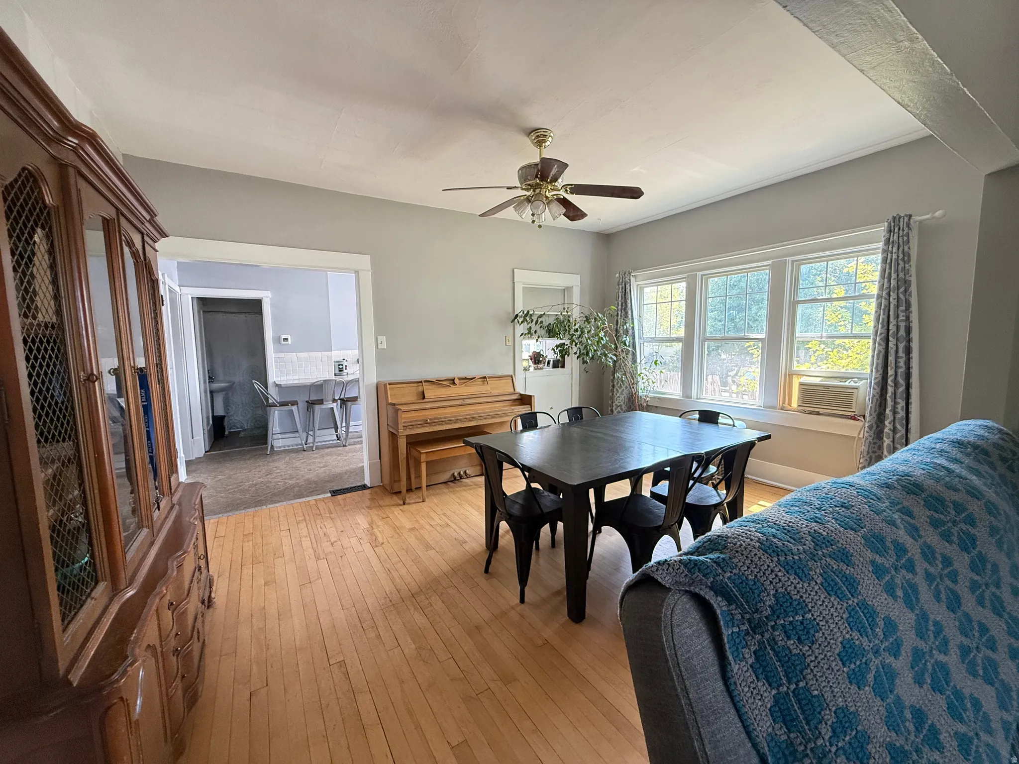 Spacious living area showcasing wood-finish flooring, a black dining table with seating for six, an upright piano, ceiling fan with integrated lighting, and abundant natural light from multi-pane glass doors and windows
