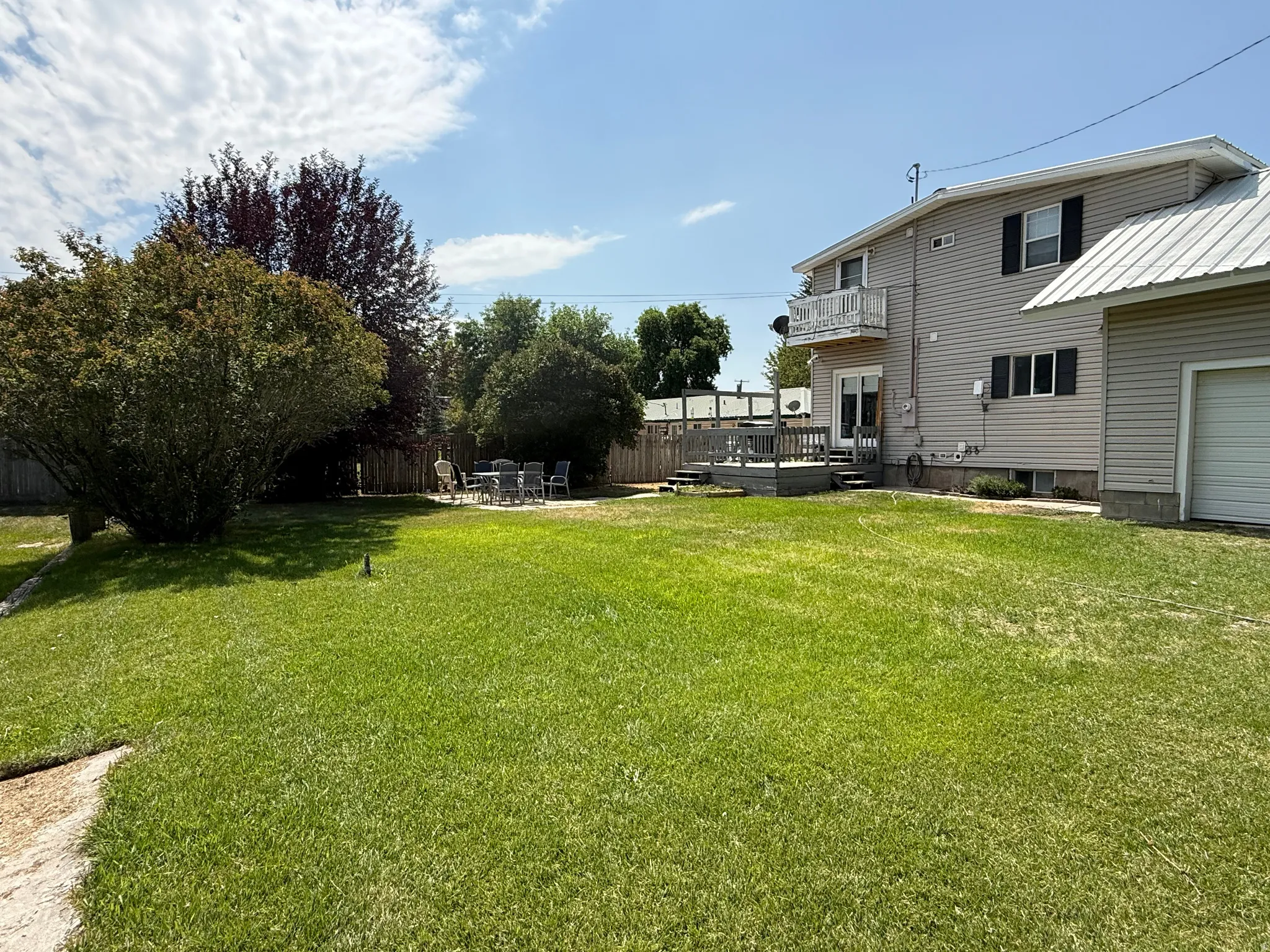 Expansive manicured lawn with mature landscaping and established trees, complemented by a two-story residence featuring metal roof accents, vinyl siding, and a multi-level deck with railings