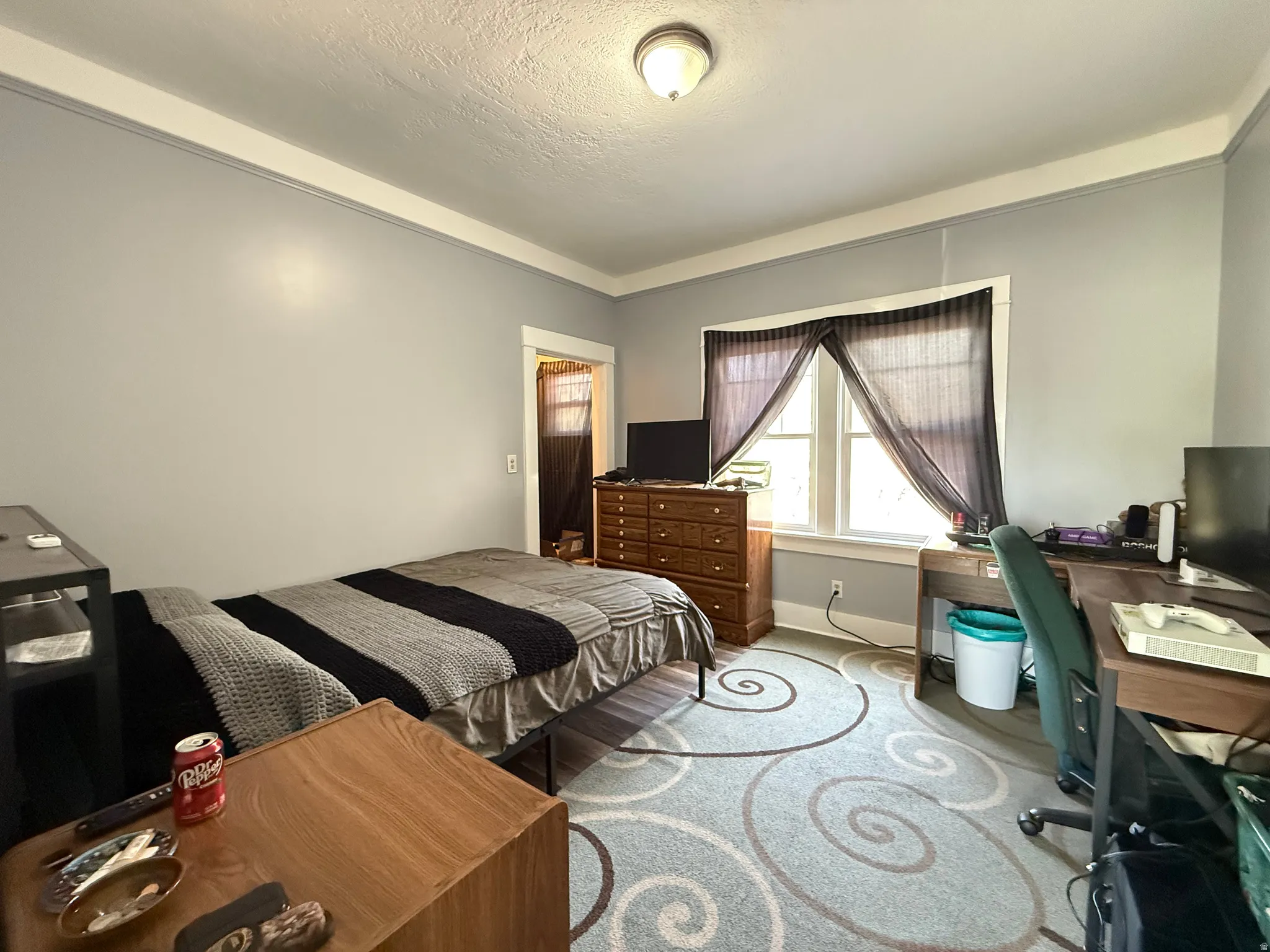 Spacious bedroom featuring wood-finish flooring, a patterned area rug, large windows with black-framed barn-style shutters and sheer curtains, wood dresser with ample storage, built-in desk area with shelving, and recessed ceiling lighting