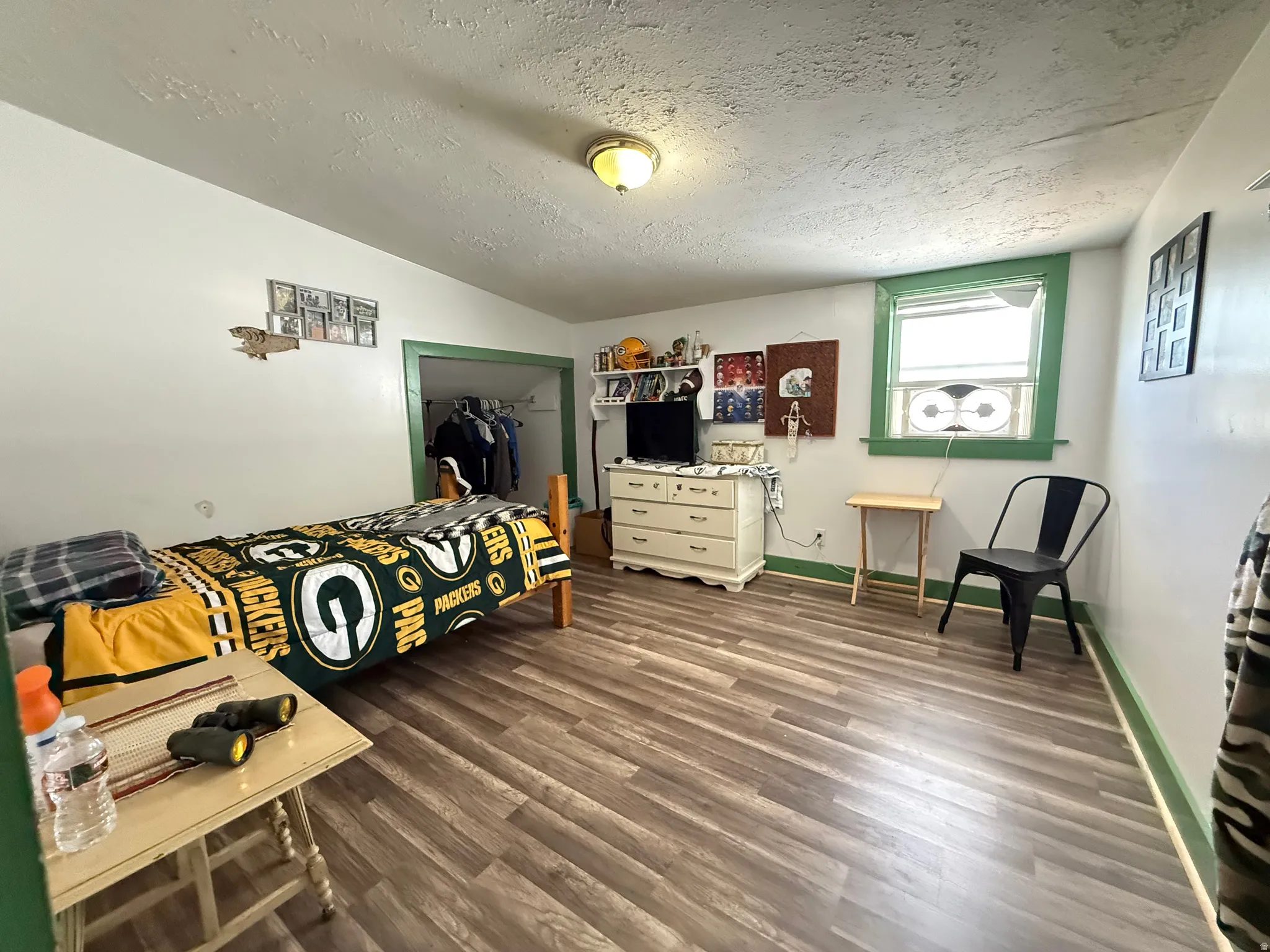Spacious bedroom with wood-finish flooring, large operable window with green trim providing natural light, built-in dresser with ample storage, ceiling-mounted fixture, and generous square footage ideal for furnishing flexibility