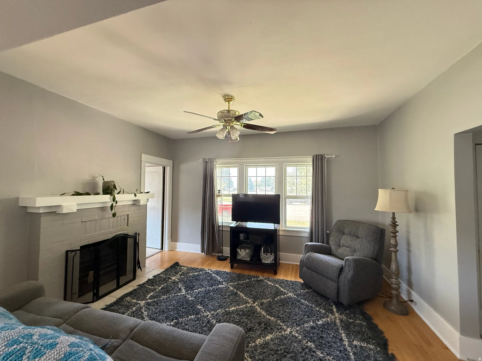 Bright living room featuring hardwood flooring, a ceiling fan with integrated lighting, a fireplace with black surround, large multi-pane windows with thermal curtains, and a dark wood entertainment console