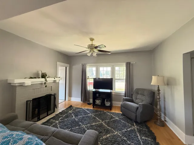 Bright living room featuring hardwood flooring, a ceiling fan with integrated lighting, a fireplace with black surround, large multi-pane windows with thermal curtains, and a dark wood entertainment console