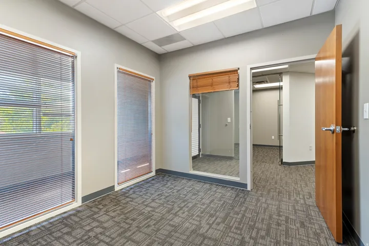 Bright, open office space featuring recessed ceiling lighting, large glass-panel interior windows with wood-finish framing, neutral wall finishes, and durable patterned carpet flooring
