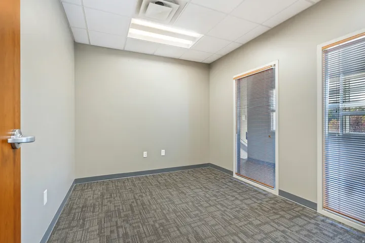 Bright, open office space featuring neutral wall finishes, recessed ceiling lighting, patterned carpet flooring, and dual glass-panel doors with aluminum framing
