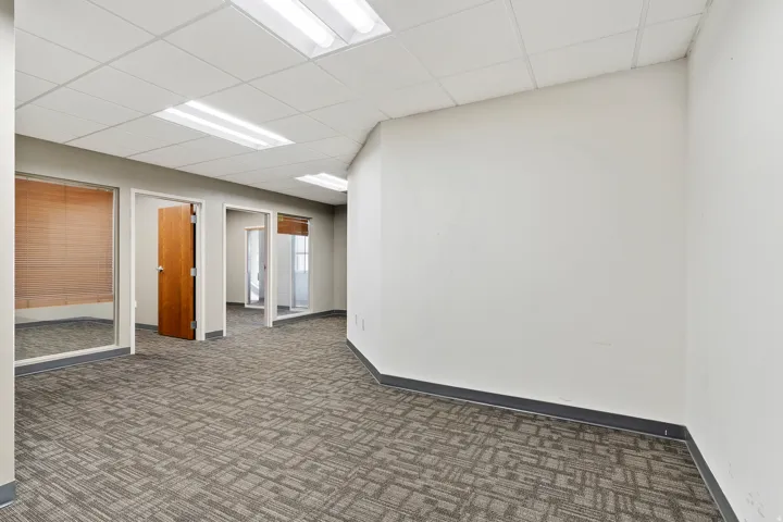 Open-plan space featuring patterned carpet flooring, white walls, drop ceiling, and multiple glass-partitioned rooms with wood doors