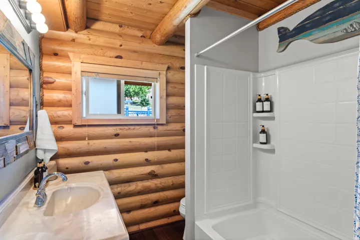 Rustic log cabin bathroom featuring stacked timber walls with exposed beam ceiling, dual vanity with white countertop and chrome faucetry, white subway tile shower enclosure with built-in shelving, and a window framing natural light and outdoor scenery