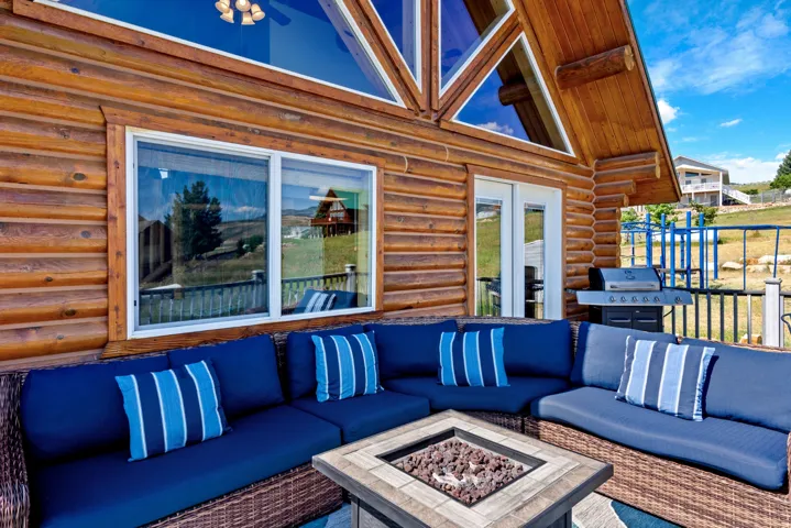 Expansive covered deck featuring custom blue upholstered seating with striped accent pillows, brick-lined fire pit with stone surround, and large multi-pane windows framing water and mountain vistas