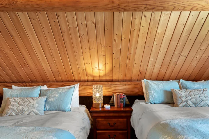 Cozy bedroom retreat featuring warm wood-finish vaulted ceiling with integrated recessed lighting, two queen beds with coordinating bedding, and built-in wooden nightstand with drawer storage between sleeping areas