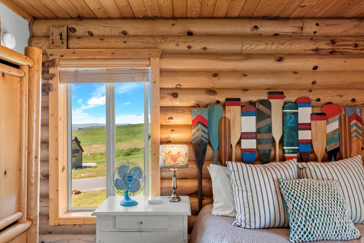 Rustic log cabin bedroom featuring exposed timber beam construction with natural wood detailing, large multi-pane windows framing expansive pastoral landscape, built-in bed frame with striped bedding, and integrated wall-mounted lighting fixtures