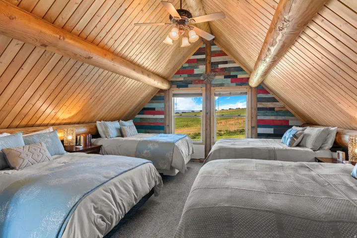 Spacious loft bedroom with soaring wood-beamed ceilings, multiple beds, and abundant natural light from large dual windows framing pastoral landscape views