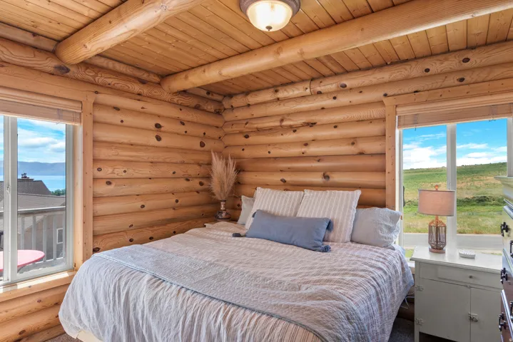 Bright bedroom retreat showcasing handcrafted log construction with exposed beam ceiling, large multi-pane windows framing pastoral landscape vistas, and soft natural light throughout