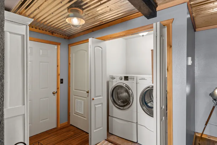 Dedicated laundry room featuring side-by-side front-load washers and matching pedestals, wood-finish beams and trim, corrugated metal ceiling detail, white six-panel doors, and wood-finish flooring throughout