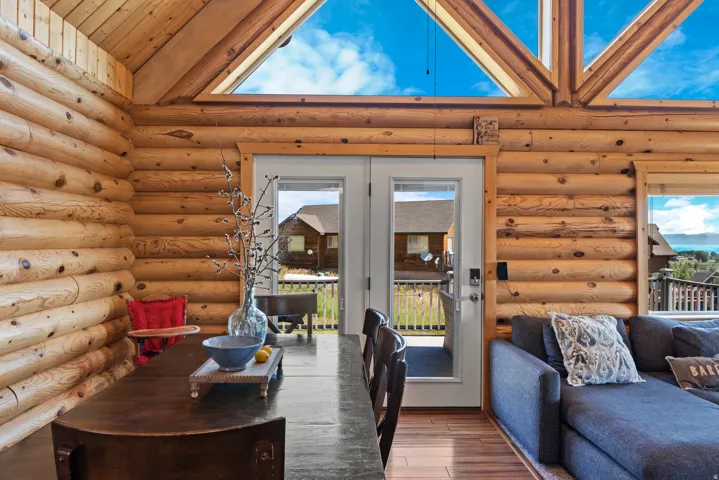 Rustic log cabin interior showcasing soaring cathedral ceilings with exposed beam trusses, expansive glazed doors and transom windows flooding the space with natural light, wood-finish flooring, and a rounded wood-finish dining table