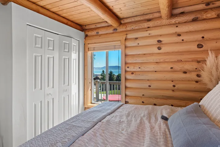 Bright bedroom featuring rustic log construction with exposed timber beams and ceiling, spacious closet with six-panel white doors, and glass sliding doors opening to a covered deck with scenic outdoor vistas