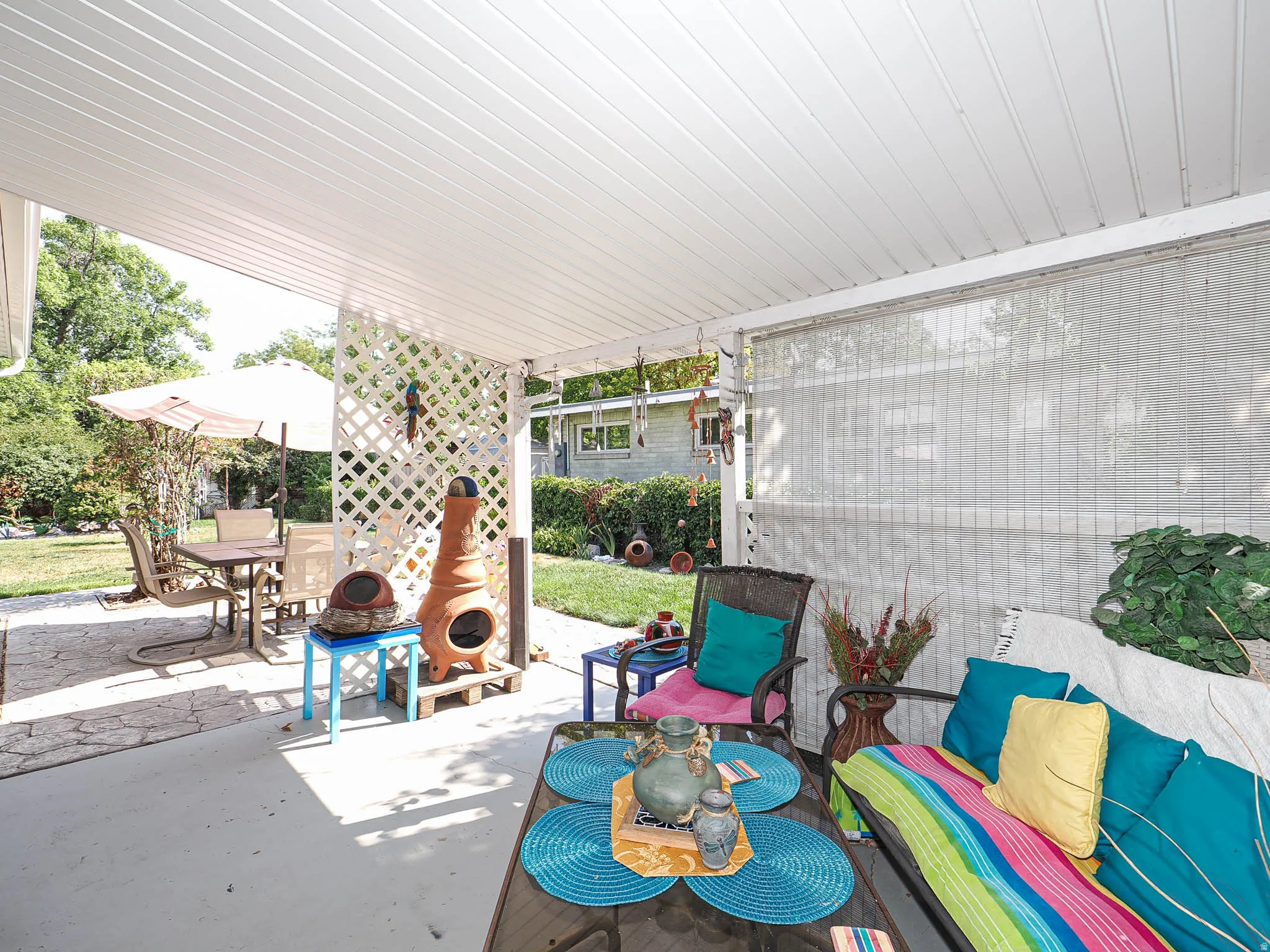 Covered patio with white beadboard ceiling and ceiling fan, featuring a blue tile-top bistro table, built-in bench seating with colorful cushions, and access to the fenced yard beyond