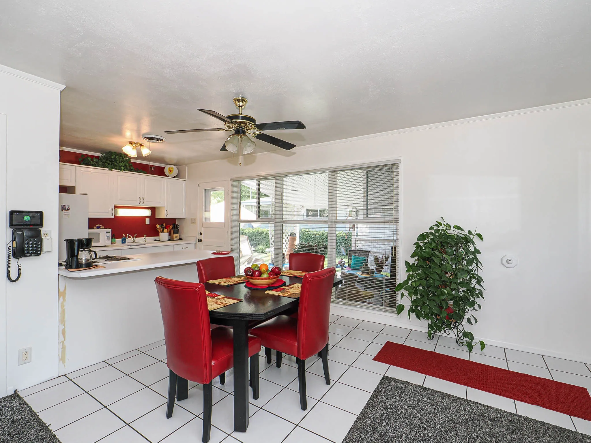 Bright, open kitchen and dining area featuring tile flooring, stainless steel appliances, red upholstered dining chairs, ceiling fan with integrated lighting, and sliding glass doors providing natural light and access to the outdoor space