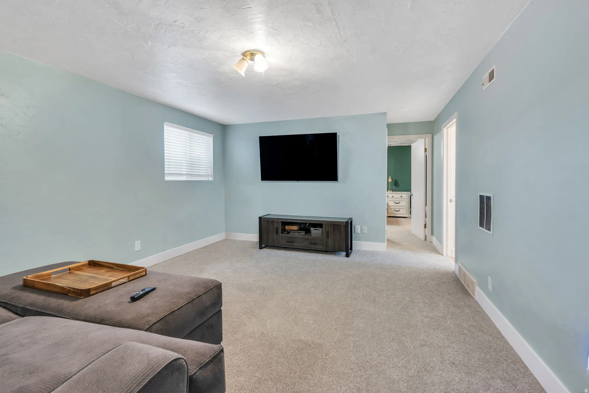 Spacious recreation room featuring light blue painted walls, neutral carpet flooring, a wall-mounted flat-screen television, and a built-in media console with storage