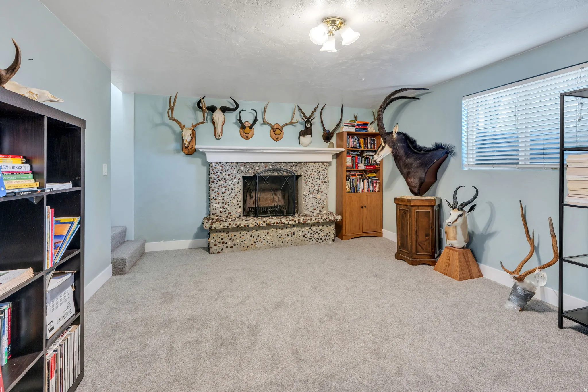 Spacious recreation room featuring a stone fireplace with wood mantel, carpeted flooring, recessed ceiling lighting, and built-in wooden cabinetry