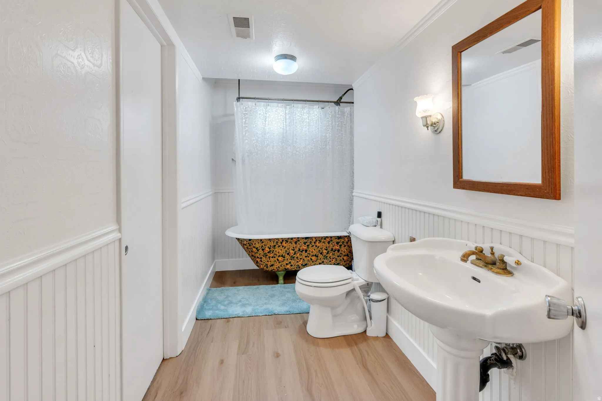 Pedestal sink, wood-finish flooring, wainscoting with chair rail, clawfoot tub, and a wood-framed mirror
