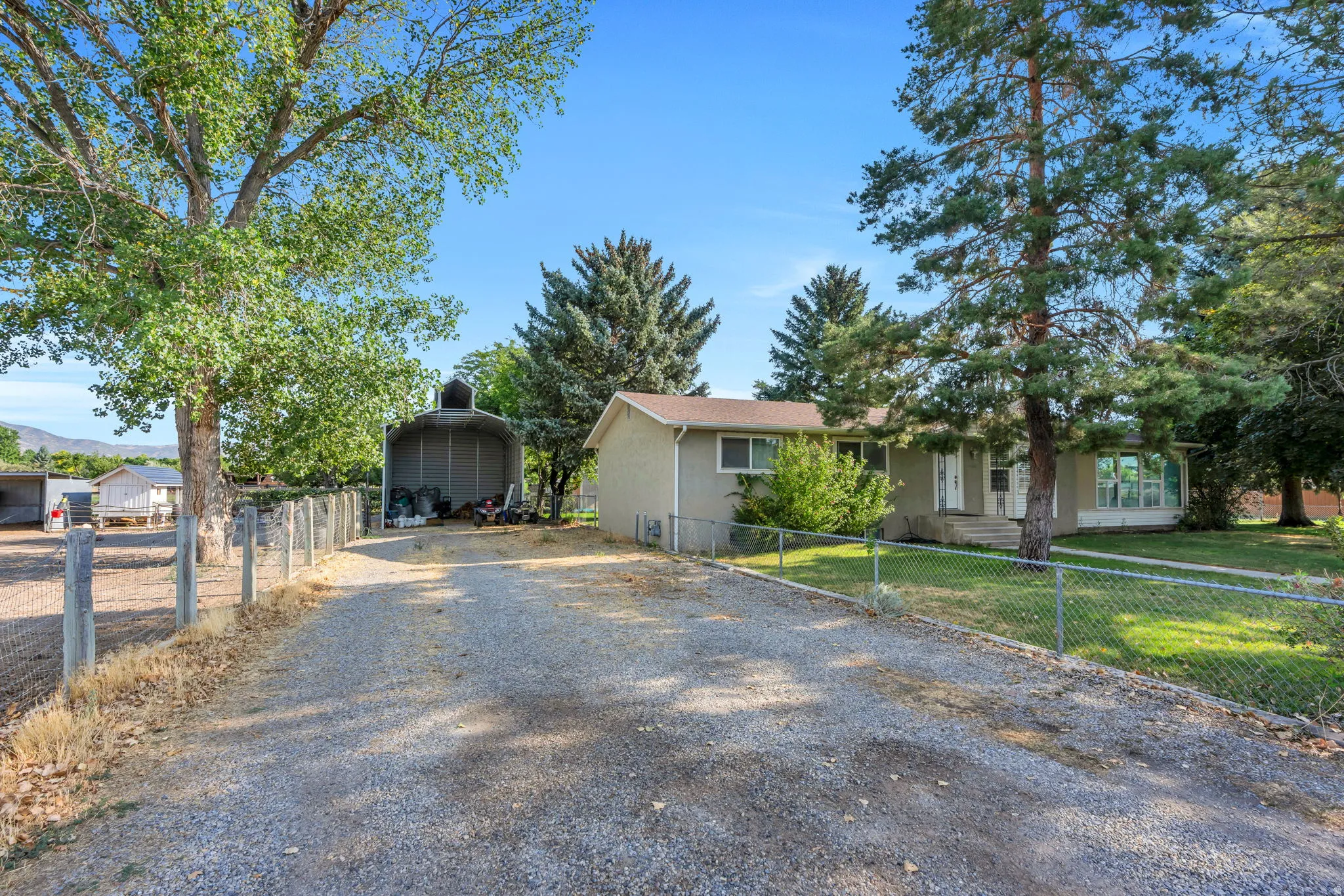 Expansive gravel driveway with mature tree-lined setting, fenced perimeter, and multiple outbuildings providing storage and utility space