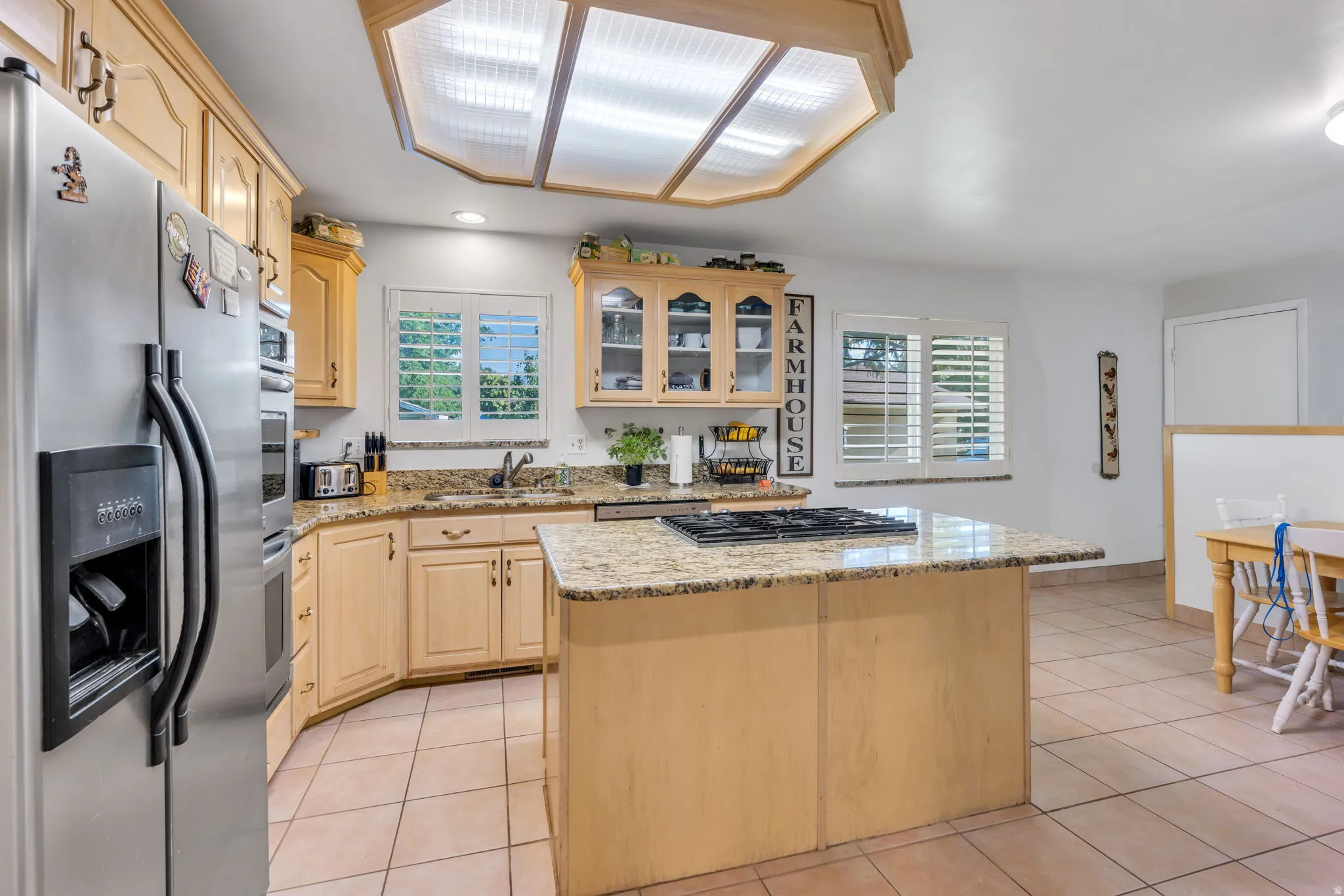 Chef's kitchen featuring custom wood cabinetry, granite countertops, a large center island with prep space, stainless steel appliances including a side-by-side refrigerator, and abundant natural light from a skylight and multiple windows with plantation s