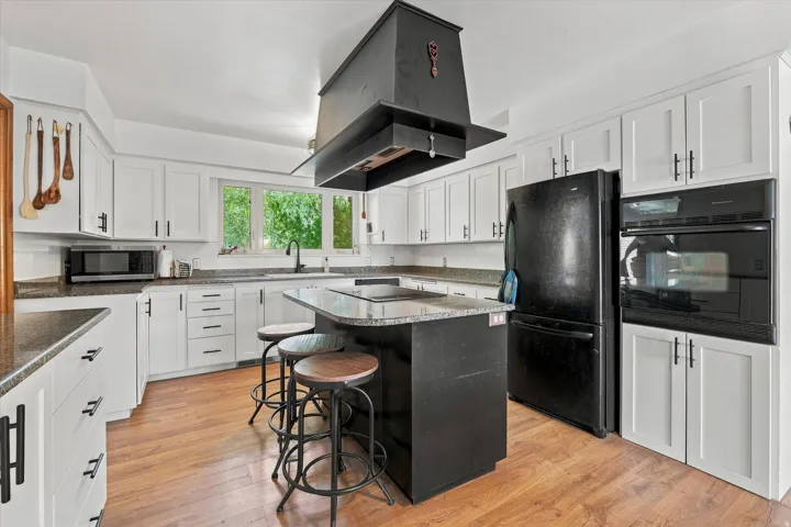 Modern kitchen featuring white shaker-style cabinetry, black stainless steel appliances, a large center island with seating, quartz countertops, wood-finish flooring, and a black ceiling-mounted range hood