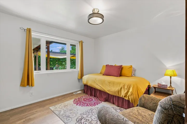 Bright secondary bedroom featuring hardwood flooring, ample natural light from multi-pane windows with gold curtain hardware, and a ceiling-mounted light fixture