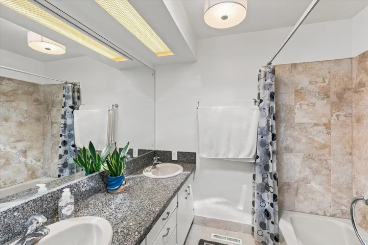 Modern bathroom featuring dual vanity with speckled stone countertop, recessed ceiling lighting, tile surround in neutral tones, and a separate shower enclosure with coordinating tile work and glass partition