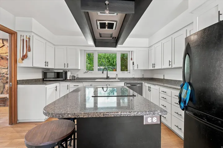 Modern kitchen featuring white cabinetry with brushed nickel hardware, a gray speckled stone island countertop with seating, stainless steel appliances including a black refrigerator, a window-lined sink area with natural light, and wood-finish flooring t