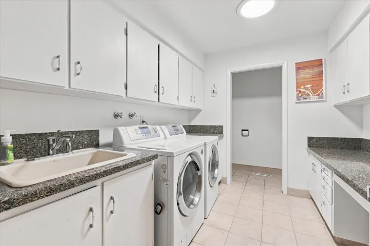 Dedicated laundry room featuring dual front-load washers and dryers, speckled stone-finish countertops, white cabinetry with ample storage, tile flooring, and a convenient pass-through to adjacent spaces