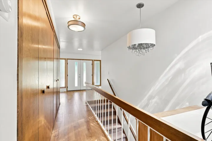 Elegant upper-level hallway featuring warm wood-finish flooring, custom wood railing with metal balusters, and abundant natural light from recessed ceiling fixtures and a decorative pendant chandelier