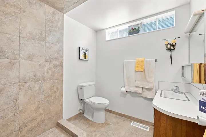 Spacious bathroom featuring ceramic tile shower enclosure with neutral stone-finish walls, white subway tile surround, pedestal sink with chrome faucet, and toilet with dual towel bars