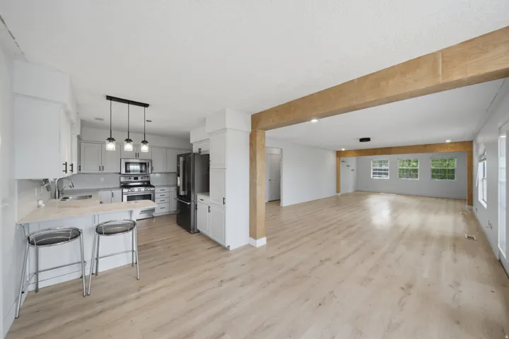 Open-concept living space with light wood-finish flooring, white walls, and an exposed wood-finish beam
