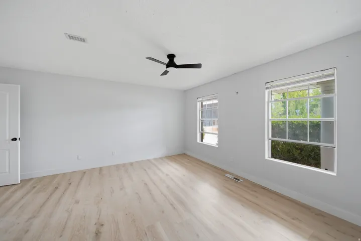 Bright, open room featuring gleaming wood-finish flooring, abundant natural light from dual large windows, a ceiling fan with integrated lighting, and clean white walls